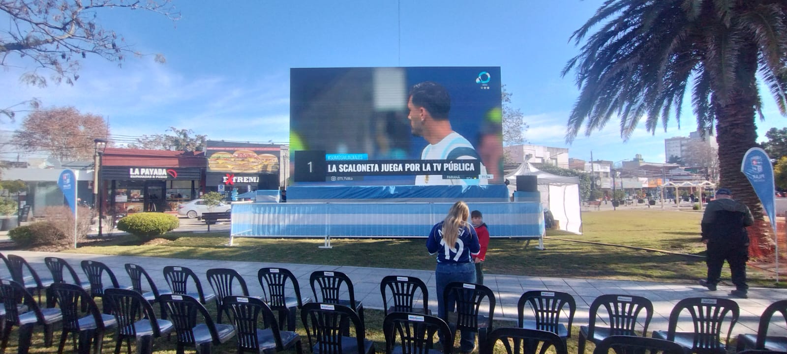 Painel LED ligado sobre gramado, com fileiras de cadeiras voltadas para a imagem.